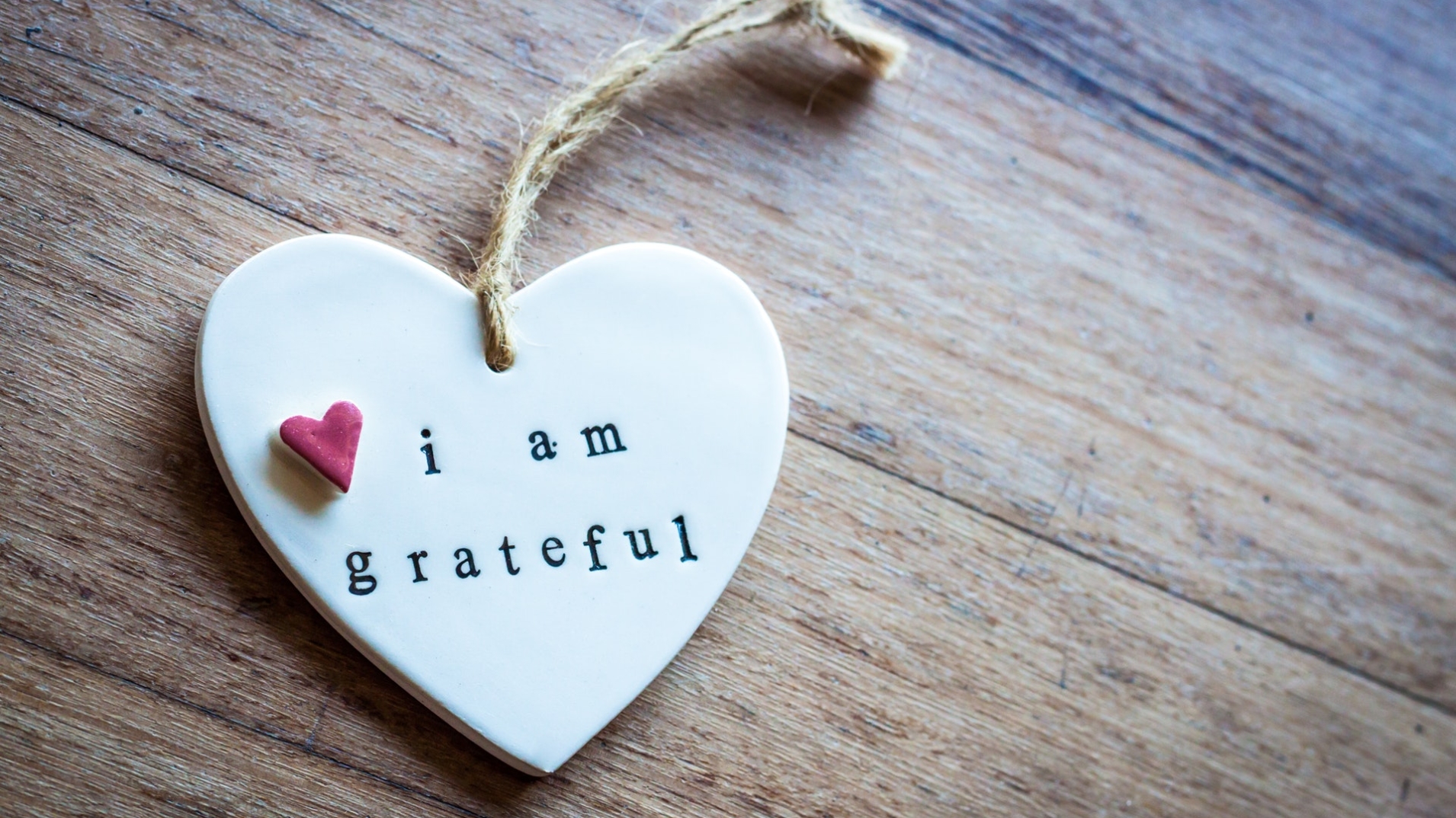 A white heart ornament is on a wooden surface. On the heart, it has a small pink heart and the words "I am grateful".